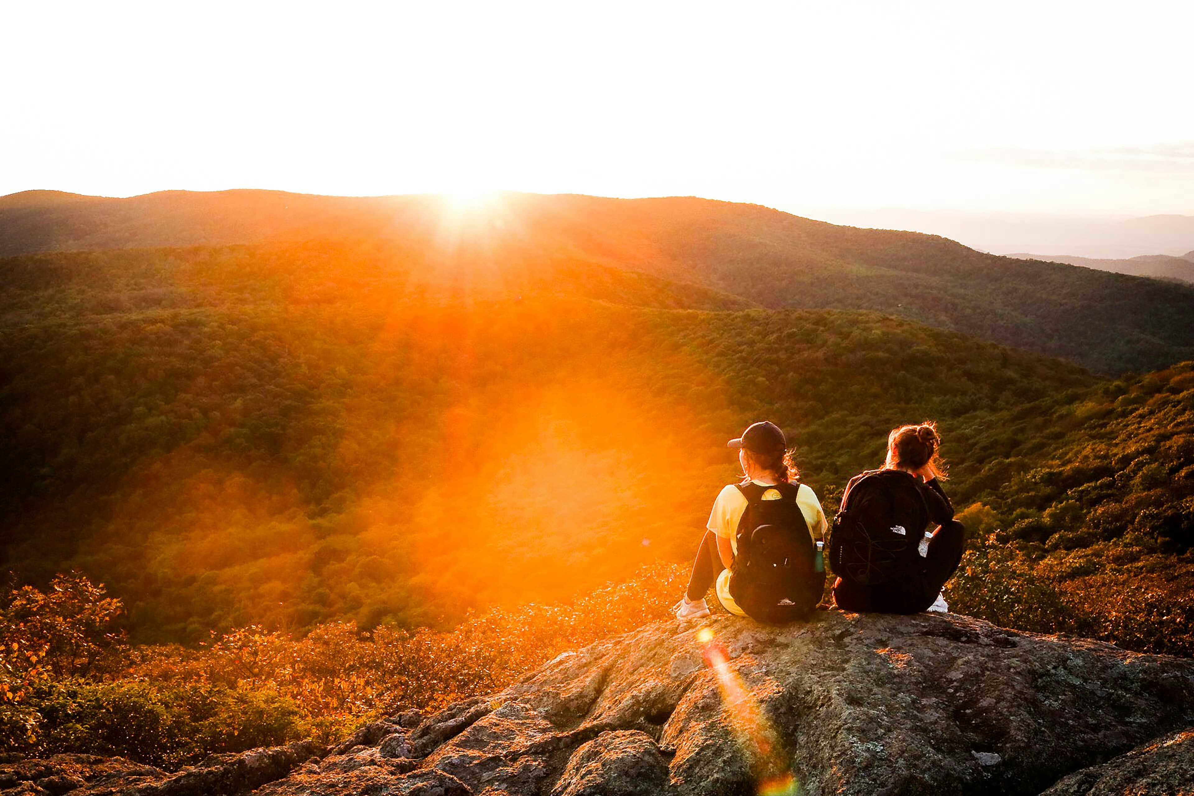 Two hikers resting on a rock outcrop at sunset over forested ridges