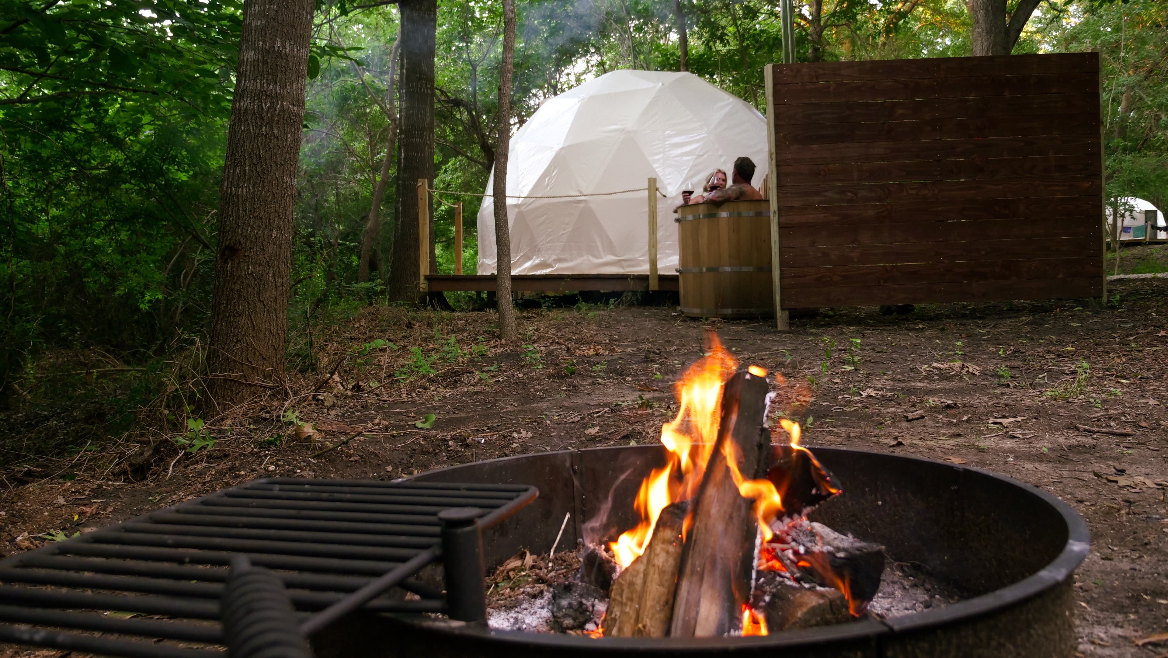 Guests in a wood-fired hot tub beside their dome, with a campfire burning in front